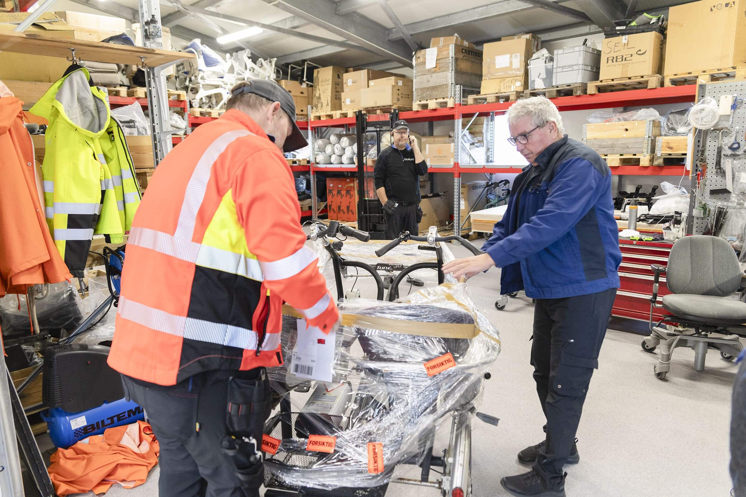Eindride Vatndal, Einar Holten og Frans Torbjørnsen pakker opp ankomne hjelpemidler som skal ut til brukere.